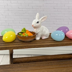 May include: A white bunny figurine sits on a wooden table with a small bowl of orange carrots, surrounded by pastel colored Easter eggs.