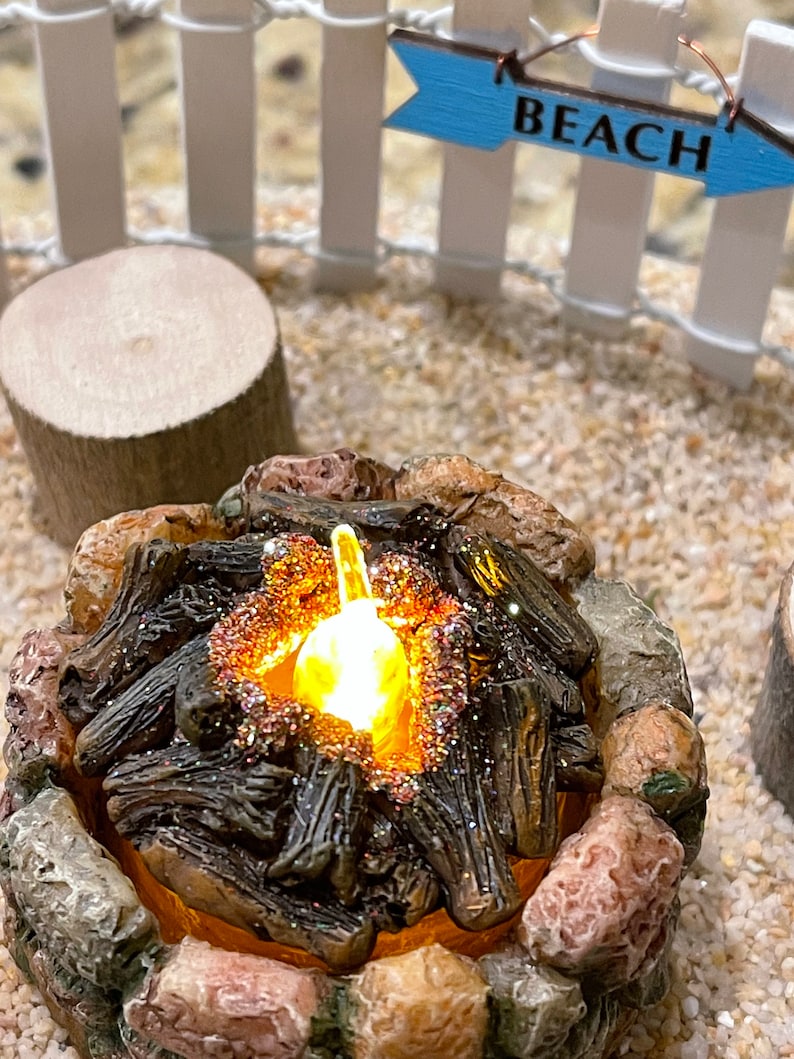 K&ouml;nnte beinhalten: Ein Miniatur-Lagerfeuer mit einem gl&uuml;henden Licht im Inneren, umgeben von Steinen und Holzscheiten. Das Lagerfeuer ist in einer Miniatur-Strandkulisse mit einem wei&szlig;en Lattenzaun und einem blauen Schild mit der Aufschrift "BEACH" aufgebaut.