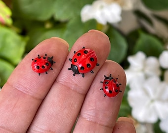 May include: Three small, red ladybug figurines with black spots and legs are displayed on a person's fingers. The ladybugs are set against a backdrop of green leaves and white flowers.