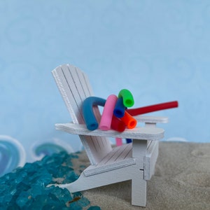 May include: A white wooden beach chair with colourful pool noodles draped over the back. The chair is sitting on a sandy beach with blue and green glass pebbles.