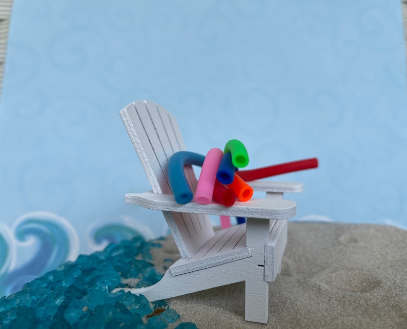 May include: A white wooden beach chair with colourful pool noodles draped over the back. The chair is sitting on a sandy beach with blue and green glass pebbles.