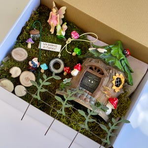 May include: A fairy garden kit in a cardboard box, featuring a miniature fairy house, figurines, mushrooms, and a sign that reads "We believe in fairies!". The scene is set on a bed of green moss, with wooden slices and decorative elements.