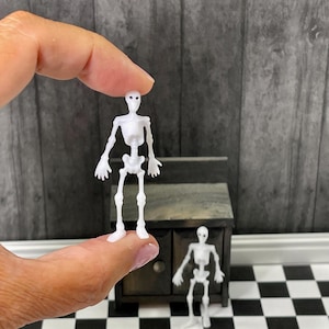 May include: Two white plastic skeleton figurines. The larger skeleton is being held by a hand. The smaller skeleton is standing on a black and white checkered floor.