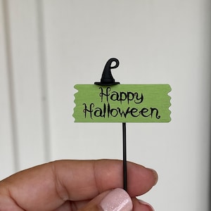 May include: A small, green sign with the words "Happy Halloween" in black script. The sign is topped with a black witch hat and mounted on a thin, black stake. The sign is held by a person.