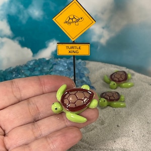May include: Miniature green and brown turtles with a turtle crossing sign. The sign is yellow with a turtle graphic and the words "TURTLE XING". The turtles are on a sandy surface.