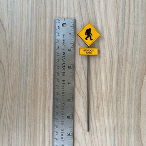 May include: A yellow warning sign with a black silhouette of a Bigfoot walking. The sign reads "Bigfoot Xing" and is attached to a black metal stick. The sign is measured against a ruler.