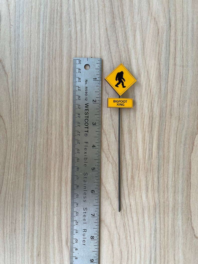 May include: A yellow warning sign with a black silhouette of a Bigfoot walking. The sign reads "Bigfoot Xing" and is attached to a black metal stick. The sign is measured against a ruler.