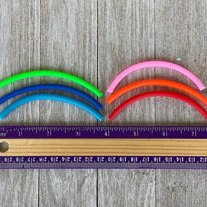 May include: Five colourful rubber bands in a rainbow pattern, measured against a ruler. The bands are green, blue, light blue, orange, and pink.