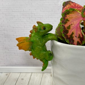 May include: A green and orange dragon figurine climbing on a white ceramic pot. The dragon has detailed scales and wings. The pot contains a plant with green, pink, and red leaves. The background is a white brick wall.