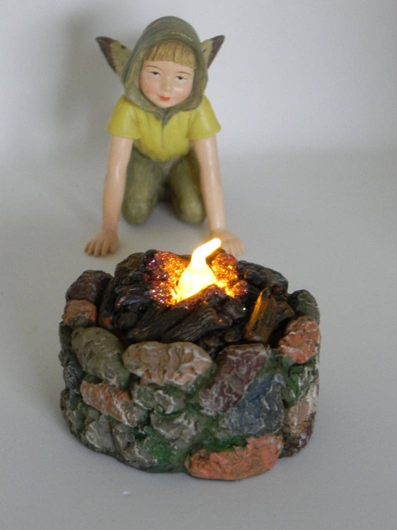 May include: A small figurine of a fairy with yellow clothing and brown wings kneeling in front of a miniature stone campfire with a glowing orange light.