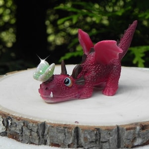 May include: A small, pink, dragon figurine with a green butterfly on its head. The dragon is lying on its belly with its wings spread out. The figurine is made of plastic and has a glossy finish.