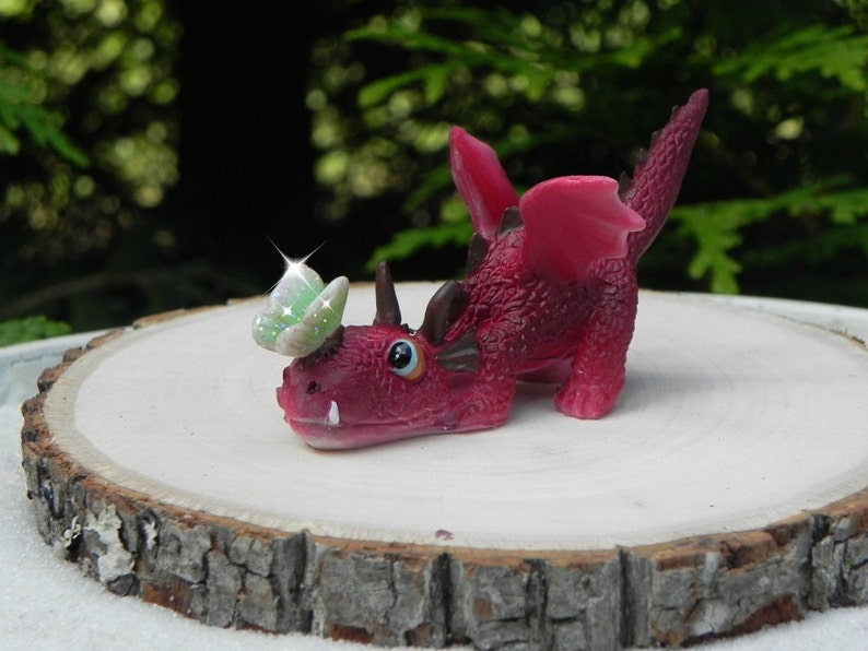 May include: A small, pink, dragon figurine with a green butterfly on its head. The dragon is lying on its belly with its wings spread out. The figurine is made of plastic and has a glossy finish.