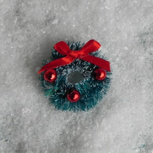 May include: A small green Christmas wreath with red berries and a red bow. The wreath is sitting on a white fluffy surface.