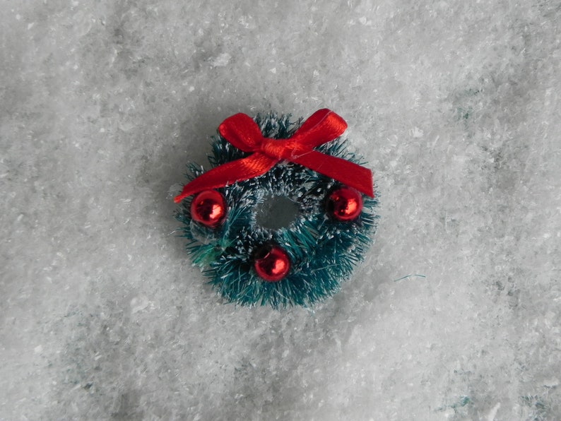 May include: A small green Christmas wreath with red berries and a red bow. The wreath is sitting on a white fluffy surface.