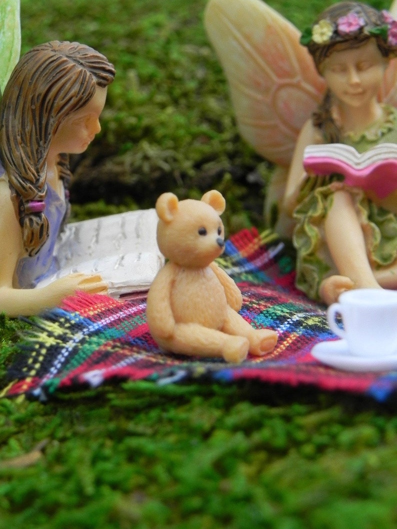 May include: A miniature scene featuring fairy figurines and a small teddy bear on a colorful plaid blanket. The fairies are reading books, and a white teacup sits nearby. The scene is set on a bed of green moss.