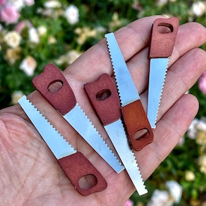 May include: A collection of five miniature hand saws featuring metallic silver serrated blades and dark reddish-brown wooden handles with a circular hole at the end of each grip, arranged in a fan-like pattern to display the tool design.