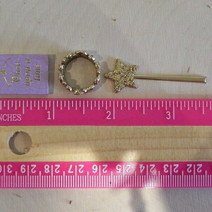 May include: A purple book with the text "Once upon a time" in gold lettering, a gold ring with a sparkly finish, and a gold star-shaped wand with a sparkly finish. The items are arranged on a wooden surface with a pink ruler measuring in inches.