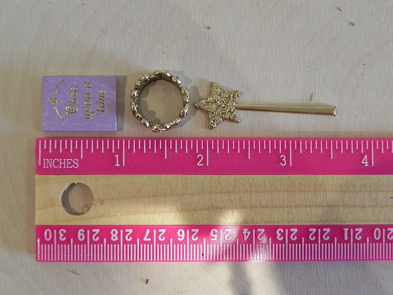May include: A purple book with the text "Once upon a time" in gold lettering, a gold ring with a sparkly finish, and a gold star-shaped wand with a sparkly finish. The items are arranged on a wooden surface with a pink ruler measuring in inches.