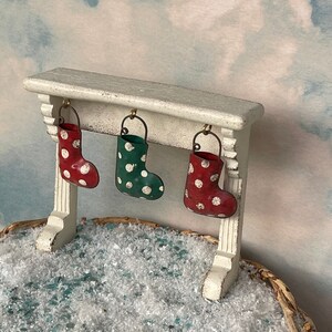 May include: A miniature Christmas scene featuring a white mantel with three hanging stockings. The stockings are red, green, and red, each adorned with white polka dots. The scene is set against a cloudy sky backdrop, with a basket filled with snow.