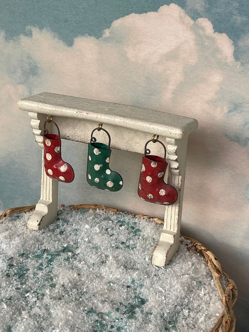 May include: A miniature Christmas scene featuring a white mantel with three hanging stockings. The stockings are red, green, and red, each adorned with white polka dots. The scene is set against a cloudy sky backdrop, with a basket filled with snow.