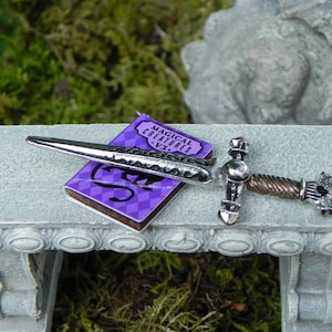 May include: A silver sword with a twisted handle and a purple book titled "Magical Creatures" on a grey stone bench.