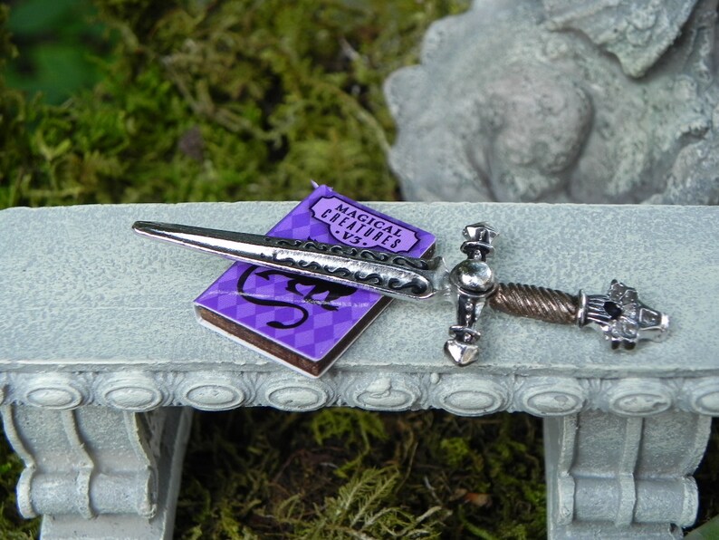 May include: A silver sword with a twisted handle and a purple book titled "Magical Creatures" on a grey stone bench.