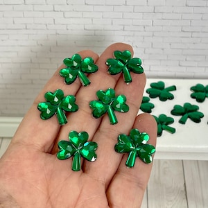 May include: A hand holding eight green shamrock-shaped rhinestones. The rhinestones are shiny and have a faceted design.