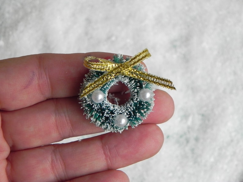 May include: A small green and white Christmas wreath with a gold ribbon and white pearl accents.