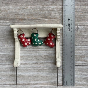 May include: A miniature Christmas mantel with three hanging stockings. Two red stockings with white polka dots flank a green stocking with white polka dots. The mantel is white with a distressed finish. A ruler shows the item is about 4 inches wide.