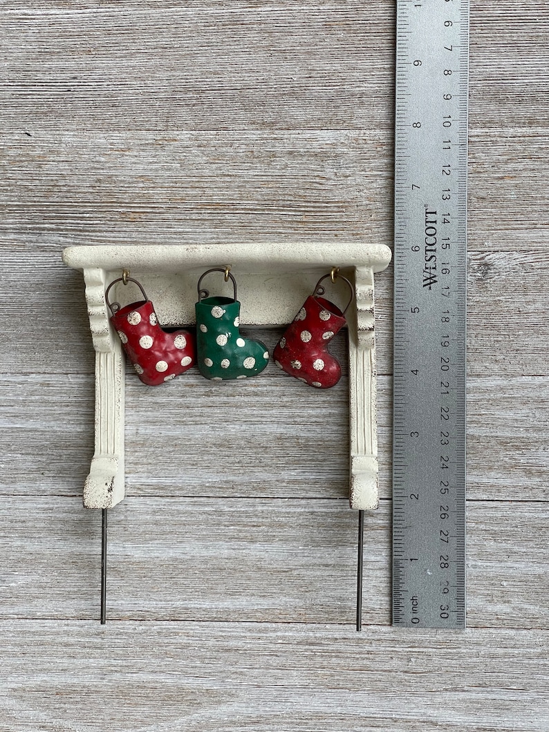May include: A miniature Christmas mantel with three hanging stockings. Two red stockings with white polka dots flank a green stocking with white polka dots. The mantel is white with a distressed finish. A ruler shows the item is about 4 inches wide.