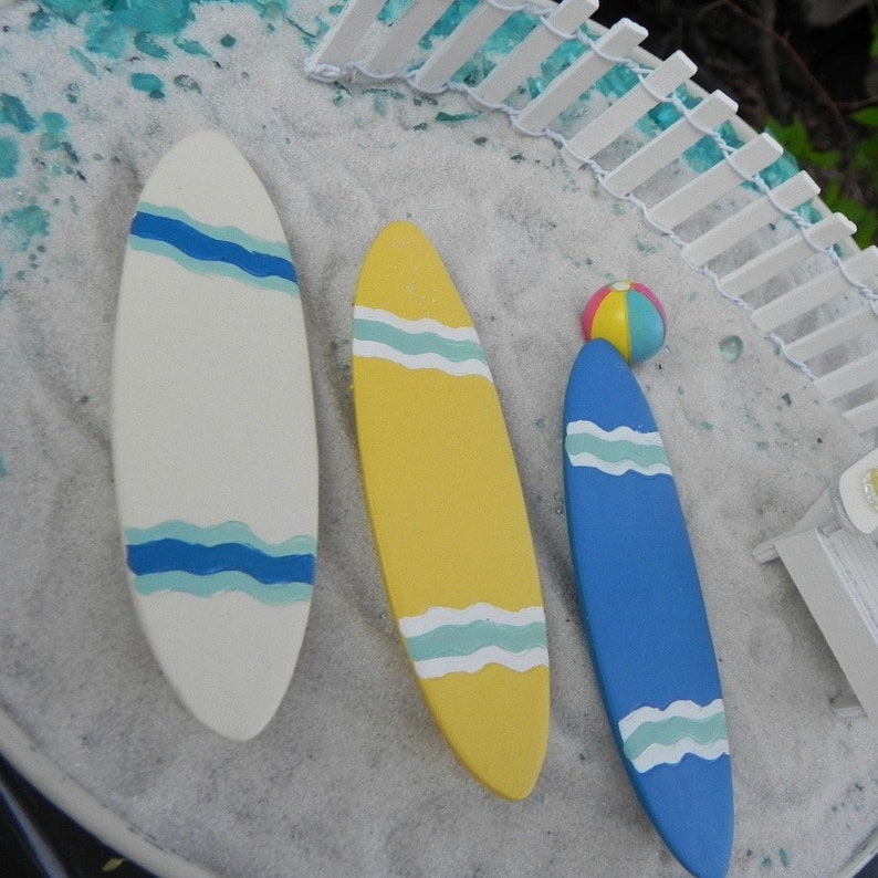 May include: Three miniature surfboards on a white sandy beach setting. The surfboards are painted in white, yellow, and blue with blue and white wave designs. A small beach ball is in the background.