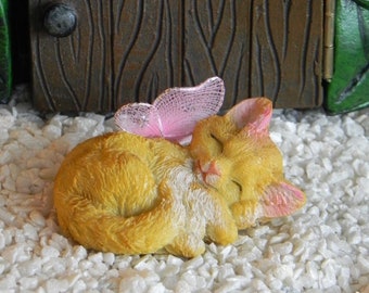 May include: A miniature figurine of a sleeping orange cat with pink-tipped ears and a pink butterfly on its back. The cat is curled up on a bed of white pebbles, with a wooden door and green foliage in the background.