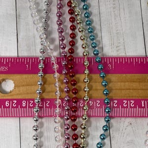 May include: Five strands of metallic bead garland in silver, clear, red, gold, and blue. The strands are laid out on a wooden ruler with pink markings.