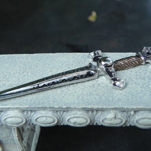 May include: A decorative silver and black sword with a detailed handle and a twisted metal design. The sword is lying on a white surface with a decorative border.