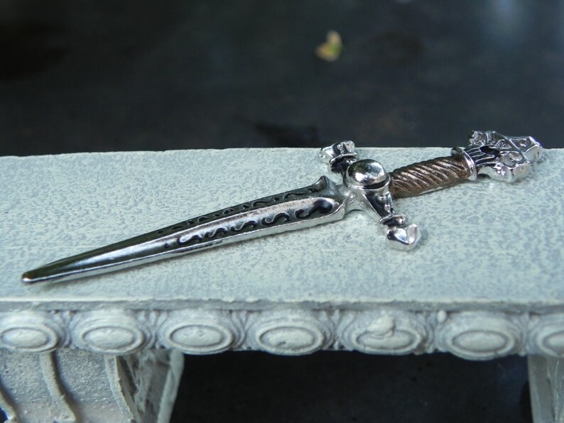 May include: A decorative silver and black sword with a detailed handle and a twisted metal design. The sword is lying on a white surface with a decorative border.