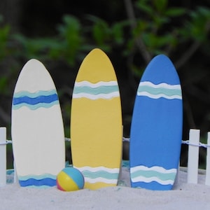 May include: Three miniature surfboards, one white with blue stripes, one yellow with blue stripes, and one blue with white and green stripes, stand in a row on a sandy surface with a white picket fence in the background. A small yellow and blue beach ball sits in front of the surfboards.