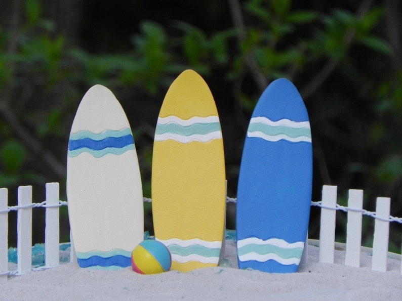 May include: Three miniature surfboards, one white with blue stripes, one yellow with blue stripes, and one blue with white and green stripes, stand in a row on a sandy surface with a white picket fence in the background. A small yellow and blue beach ball sits in front of the surfboards.
