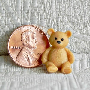 May include: A small, brown teddy bear sitting on a white textured surface. The bear is about the size of a US penny.