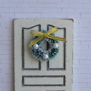 May include: A green Christmas wreath with white beads and a yellow bow hanging on a white door.