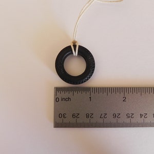 Peut inclure: Un pneu en caoutchouc noir avec une ficelle blanche attach&eacute;e, suspendu &agrave; une surface blanche. Le pneu mesure environ 2,5 cm de diam&egrave;tre. Une r&egrave;gle est visible sous le pneu, avec des marques en centim&egrave;tres.