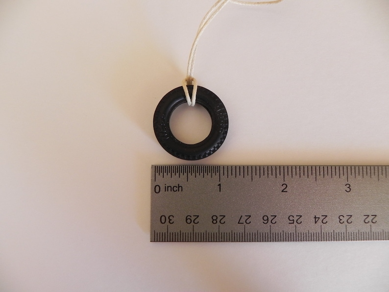 Peut inclure: Un pneu en caoutchouc noir avec une ficelle blanche attach&eacute;e, suspendu &agrave; une surface blanche. Le pneu mesure environ 2,5 cm de diam&egrave;tre. Une r&egrave;gle est visible sous le pneu, avec des marques en centim&egrave;tres.