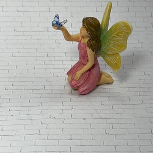 May include: A small pink fairy figurine with yellow and green wings, sitting on a white brick background. The fairy is holding a blue butterfly in her hand.