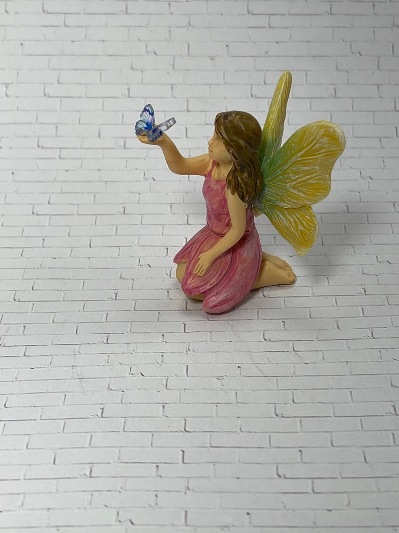 May include: A small pink fairy figurine with yellow and green wings, sitting on a white brick background. The fairy is holding a blue butterfly in her hand.