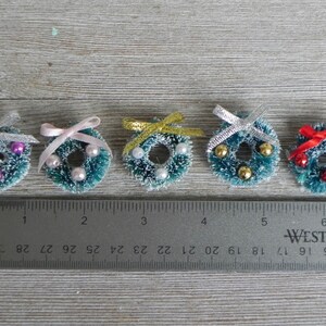 May include: Six miniature Christmas wreaths with different colored bows and ornaments. The wreaths are approximately 1 inch in diameter and are arranged in a row on a wooden surface with a ruler underneath.