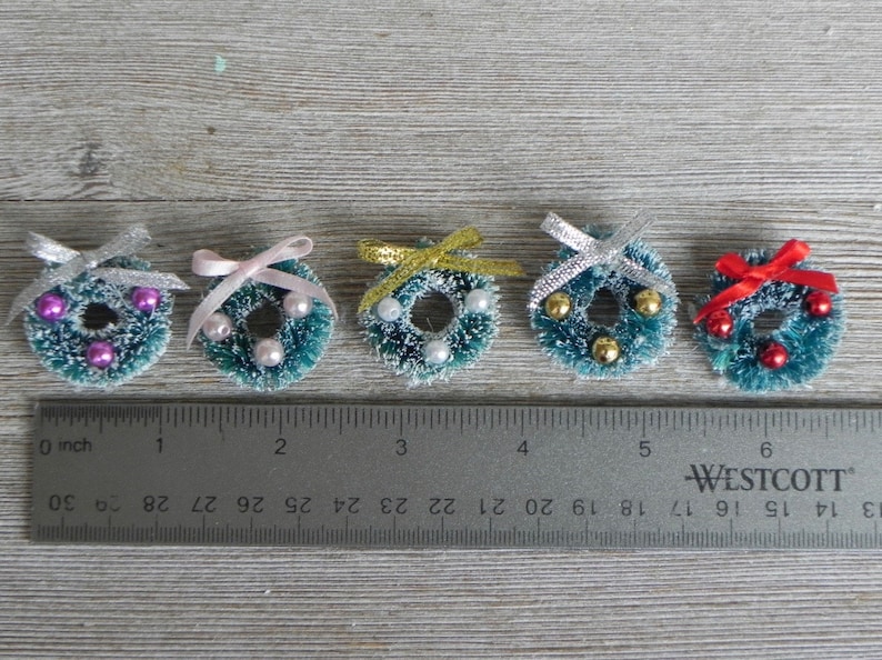 May include: Six miniature Christmas wreaths with different colored bows and ornaments. The wreaths are approximately 1 inch in diameter and are arranged in a row on a wooden surface with a ruler underneath.