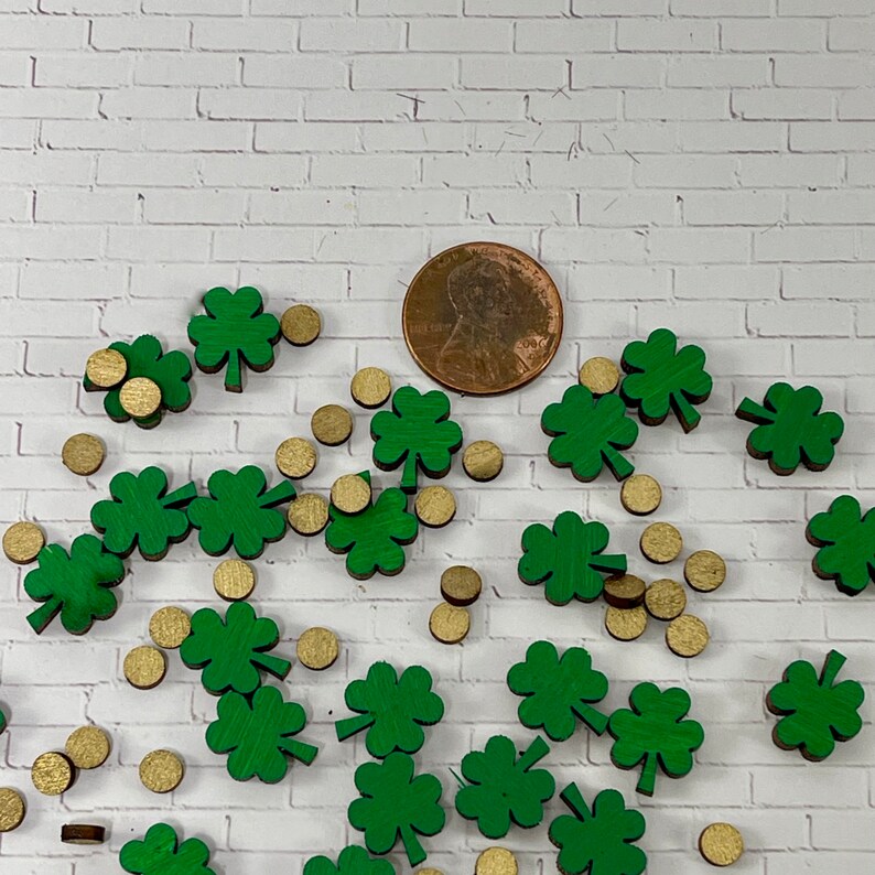 May include: A collection of green shamrock-shaped wooden cutouts and gold round wooden cutouts. The shamrocks are scattered on a white brick background.