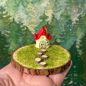 May include: A miniature fairy house with a red roof and a yellow body sits on a round wooden base covered in green moss. Small wooden stepping stones lead to the house. The base is held in a hand, with a blurred forest background.