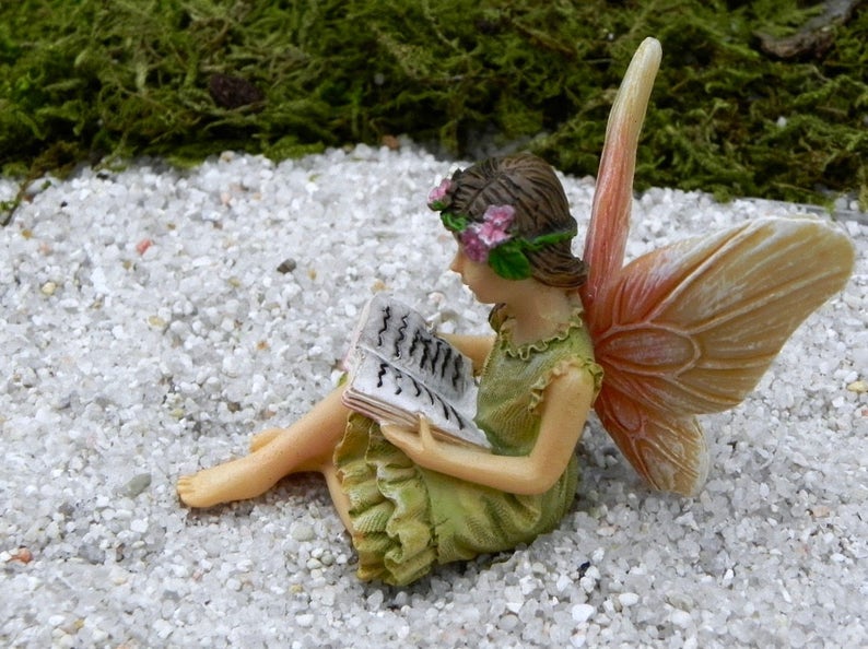 May include: A miniature fairy figurine reading a book, with a green dress and flower crown. The fairy has tan skin and large, detailed wings with peach and yellow hues. The figurine is set on a bed of white pebbles with green moss in the background.