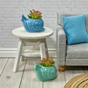 May include: Two ceramic snail planters, one light blue and one teal, each holding a small succulent plant with green and red leaves. The planters are on a white table and the floor.