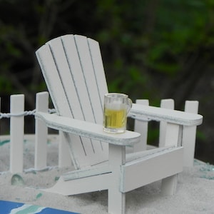 May include: A white wooden adirondack chair with a small glass of beer on the armrest. The chair is on a white sandy beach with a white picket fence in the background.